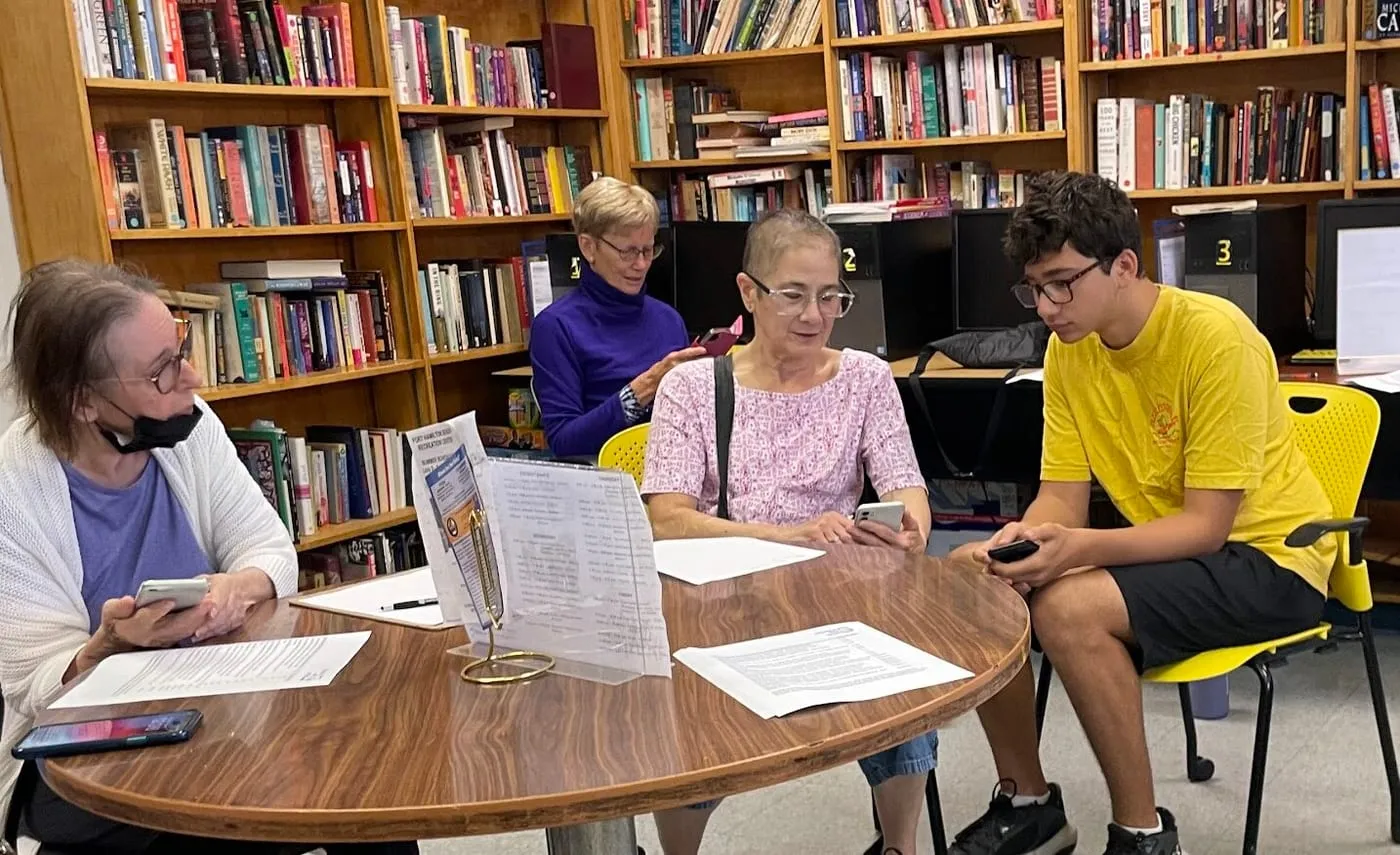 A teenage volunteer works through phone settings with three older adults around a table at the Fort Hamilton Senior Recreation Center, surrounded by library shelves.