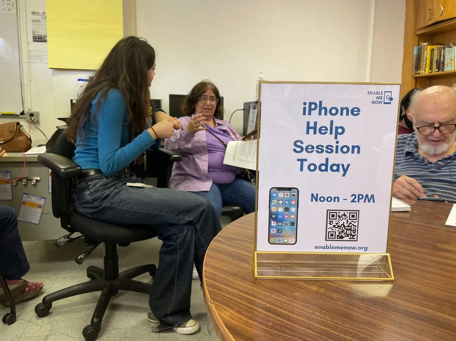 A teenage volunteer talks with a participant across a table at a community centre, beside a printed sign reading 'iPhone Help Session Today, Noon to 2PM, enablemenow.org' with a QR code.