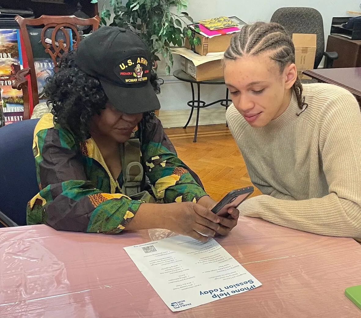 A teenage volunteer and a participant look at a phone together at a workshop table, a printed feature checklist in front of them.