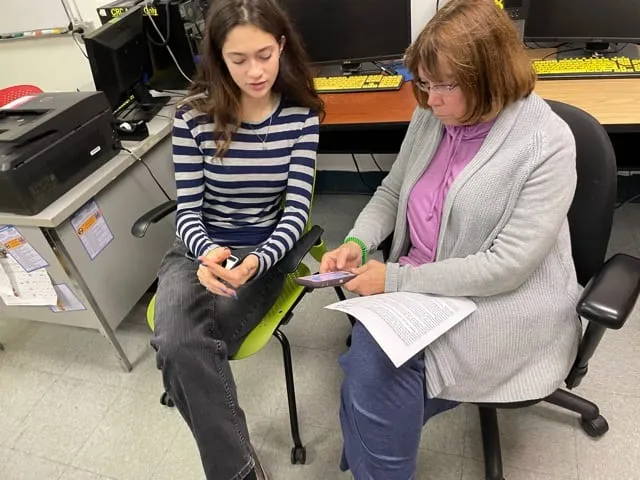 A teenage volunteer sits beside an older woman in a community-center computer room. Both are looking at her phone, and she holds a printed handout.