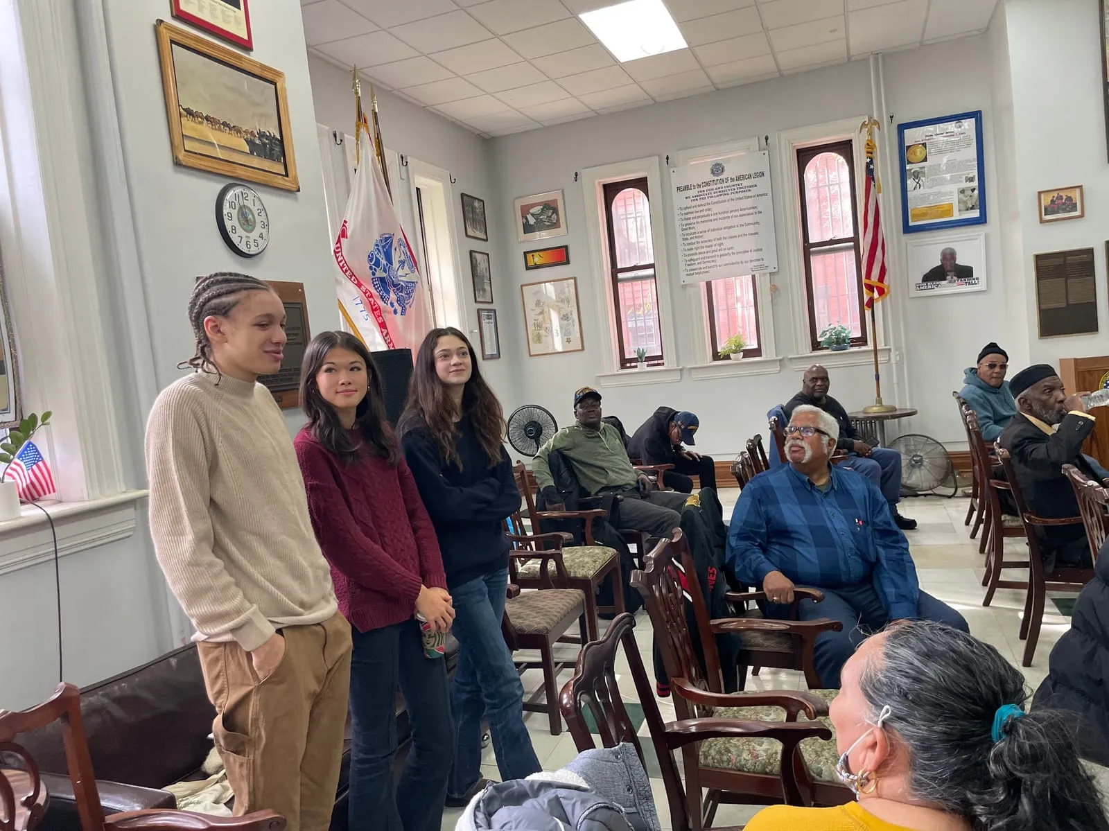 Three teenage volunteers introduce a workshop to a seated audience of older adults in a hall hung with flags and plaques.